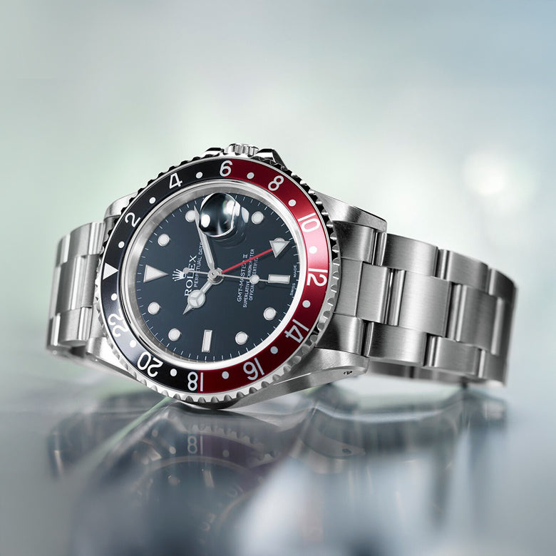 Rolex watch with a silver bracelet and black and red dial on a reflective surface.